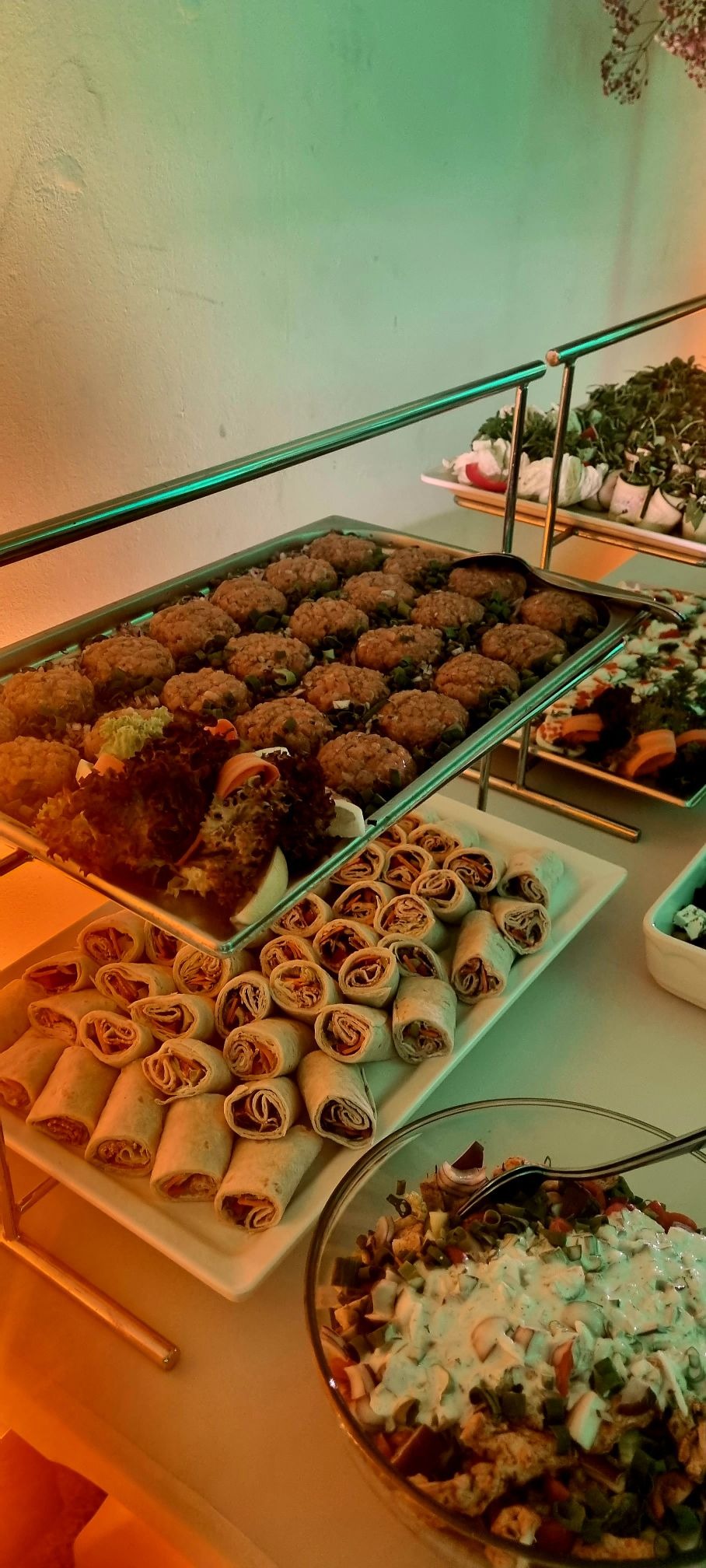 Dwupoziomowy bufet z przekąskami: roladki z tortilli, mini kotleciki z zieloną cebulką, sałatki w szklanych misach i dekoracyjne rośliny w tle, oświetlone ciepłym światłem.