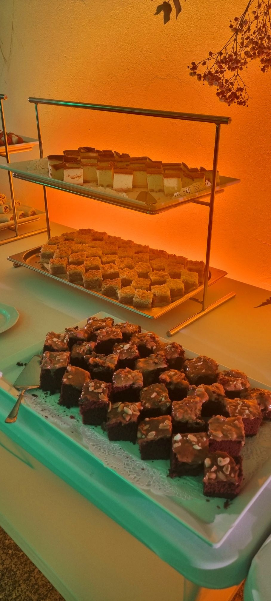 Różnorodne ciasta w kształcie kwadratów ułożone na srebrnych tacach na dwupoziomowym stojaku, oświetlone ciepłym, pomarańczowym światłem, z widocznymi drobnymi kwiatami w tle.