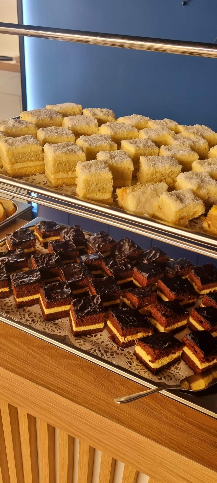 Dwa poziomy metalowej półki z kwadratowymi ciastami: na górze ciasta z kokosem, na dole ciasta czekoladowe z kremowym przełożeniem, ułożone na ozdobnej, papierowej serwetce.