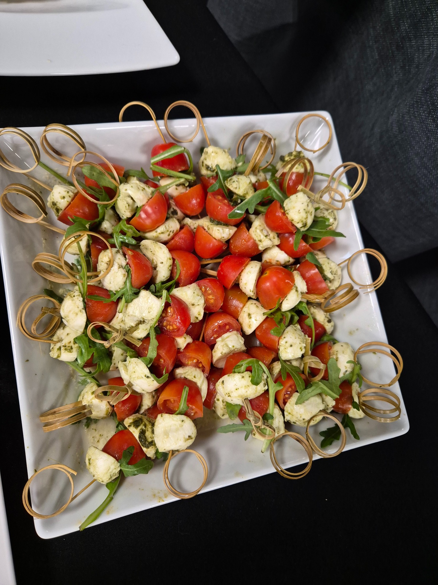 Koreczki Caprese na białym kwadratowym talerzu, składające się z pomidorków cherry, mini mozzarelli z pesto i rukoli, nabite na ozdobne bambusowe szpikulce z pętelkami.
