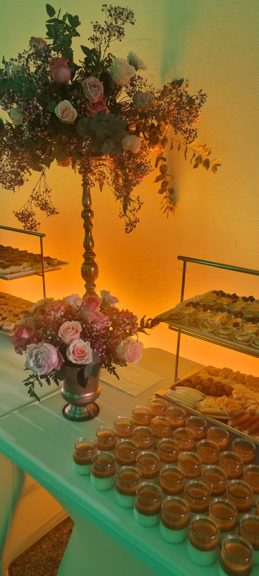 Dekoracja stołu z kwiatami w wazonie, obok desery w szklanych pojemnikach i tace z owocami i ciastami, oświetlenie w ciepłych barwach.