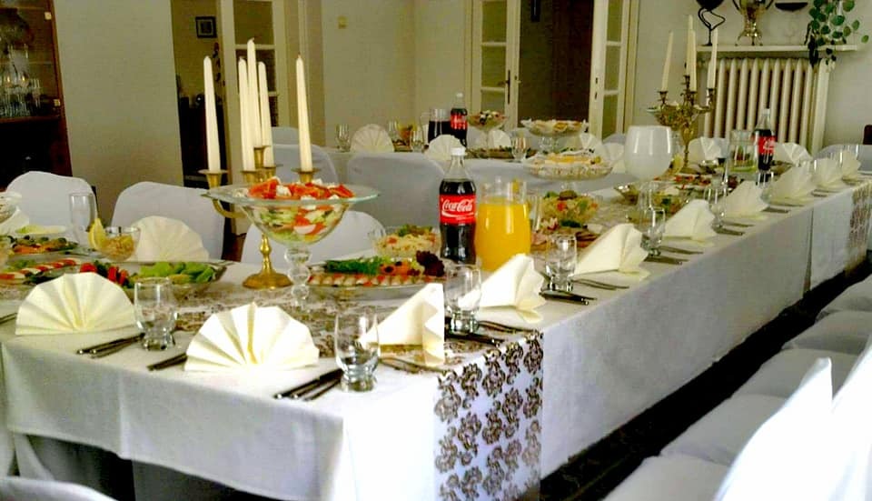 Uroczysty stół z białym obrusem, elegancko złożonymi serwetkami, szklankami, sztućcami, świecznikami z zapalonymi świecami, sałatkami w szklanych misach na nóżkach oraz napojami w butelkach i dzbanku.