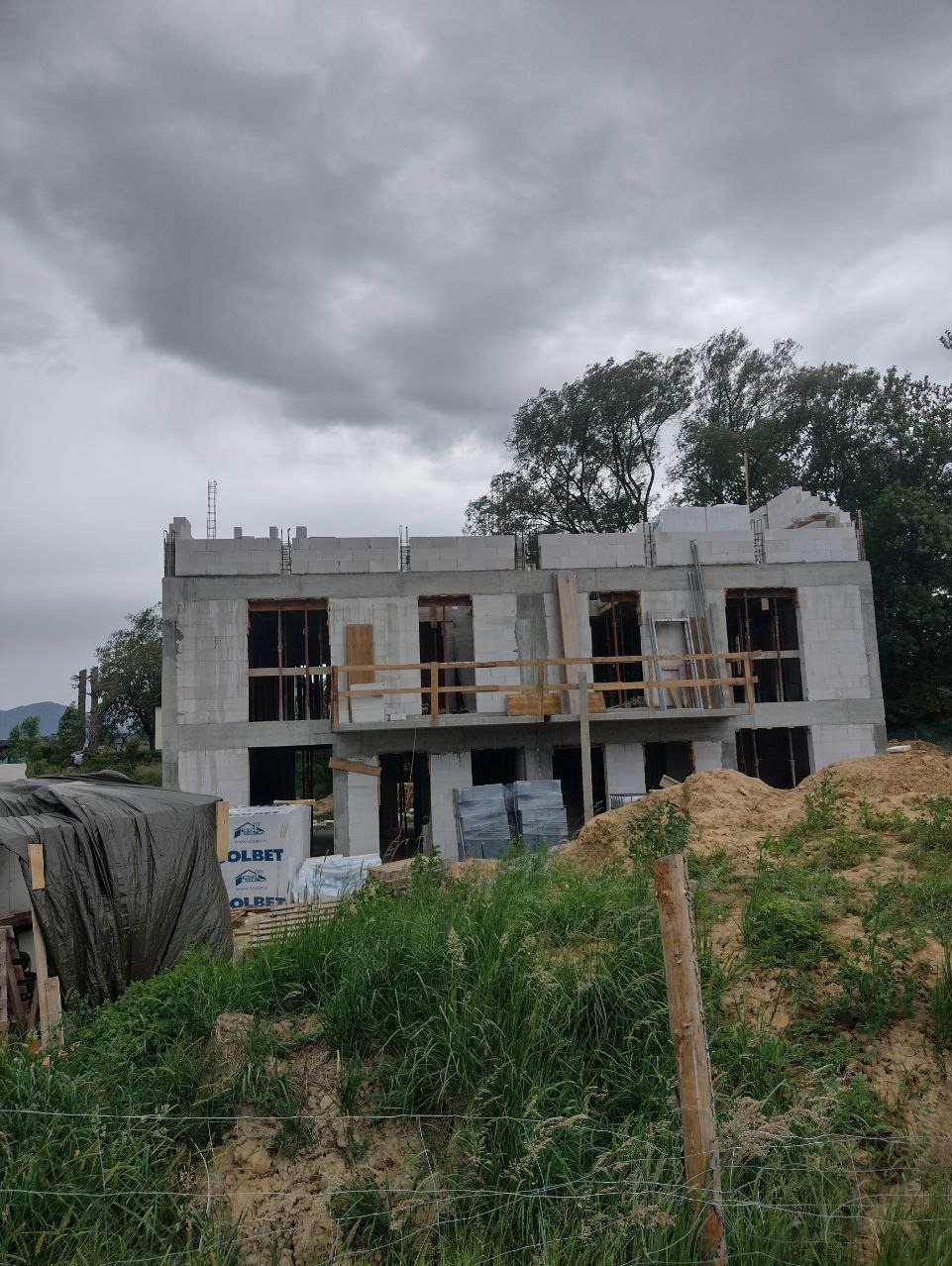 Dwukondygnacyjny budynek w trakcie wznoszenia z betonu komórkowego, widoczne niezabezpieczone otwory okienne, na pierwszym planie trawa i ziemia, w tle pochmurne niebo.