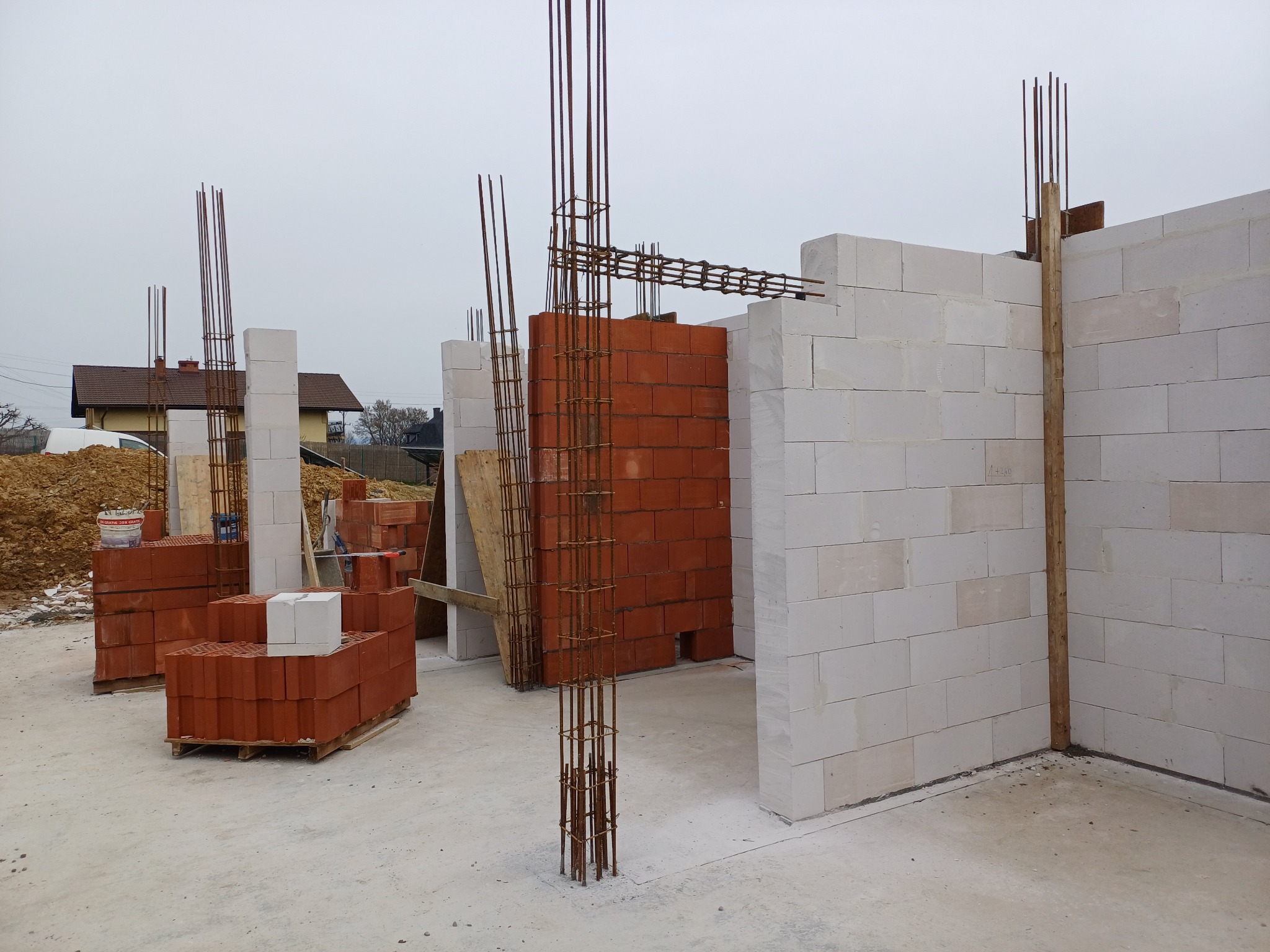 Wznoszenie ścian z bloczków białych i czerwonych, zbrojenie pionowe, widok na budowę w trakcie realizacji, w tle domy jednorodzinne.