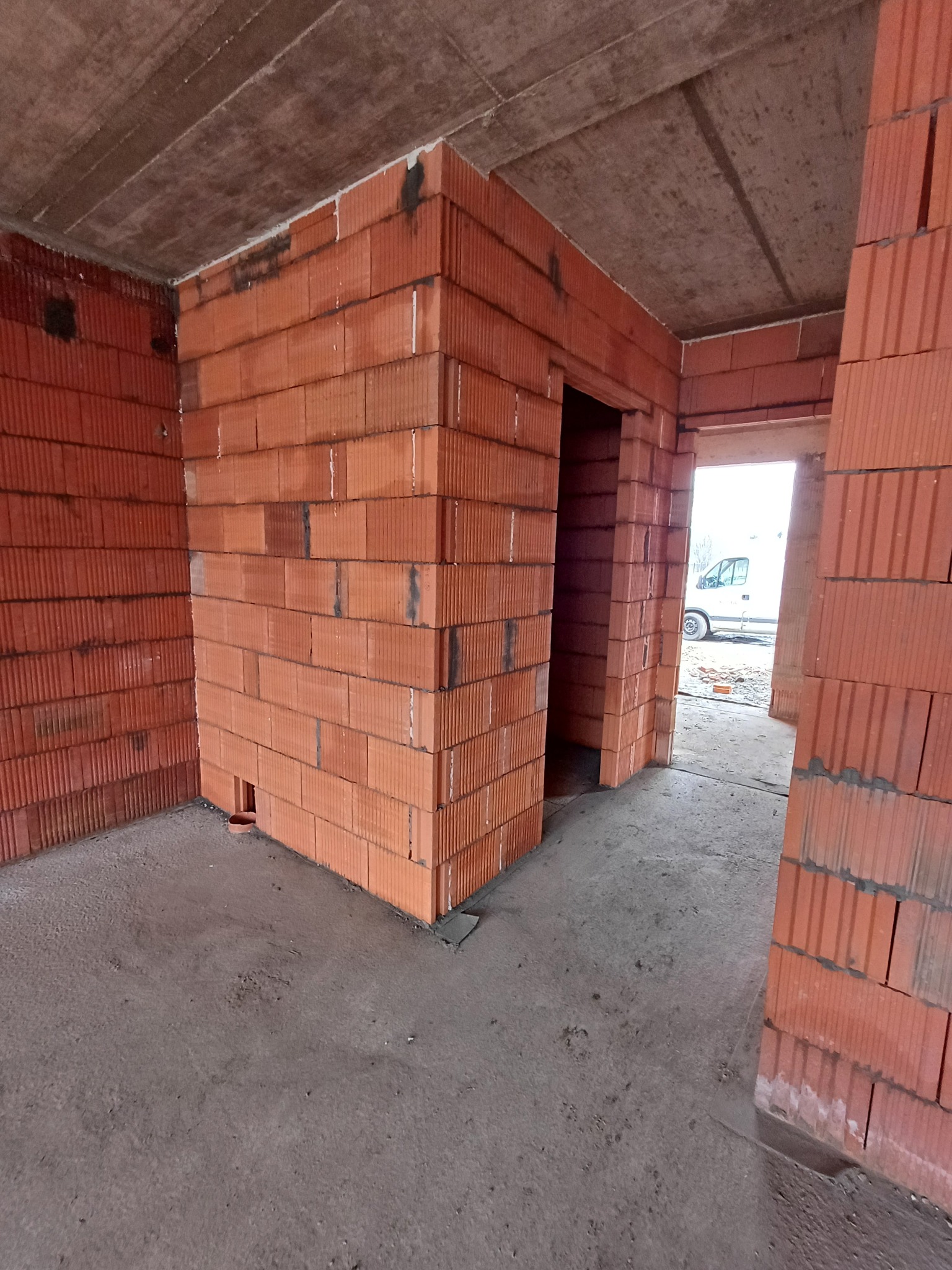 Wnętrze budynku w trakcie budowy, widoczne ściany z czerwonej cegły ceramicznej, surowy betonowy strop i betonowa podłoga, w tle przez otwór drzwiowy widoczny biały samochód dostawczy.