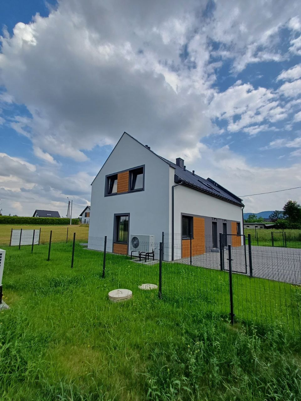 Nowoczesny dom jednorodzinny z białymi ścianami i drewnianymi akcentami, widoczny z perspektywy ogrodu z metalowym ogrodzeniem. Na elewacji zamontowana klimatyzacja, a na dachu panele słoneczne.