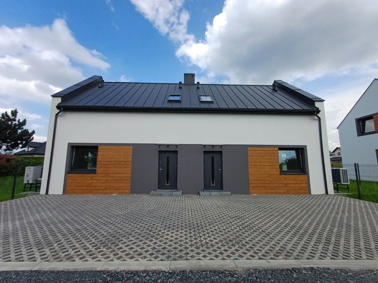 Bliźniak z białymi ścianami, grafitowym dachem i dwoma wejściami, wykończony drewnianą okładziną i grafitowymi ramami okiennymi, z ażurowym podjazdem z kostki betonowej.