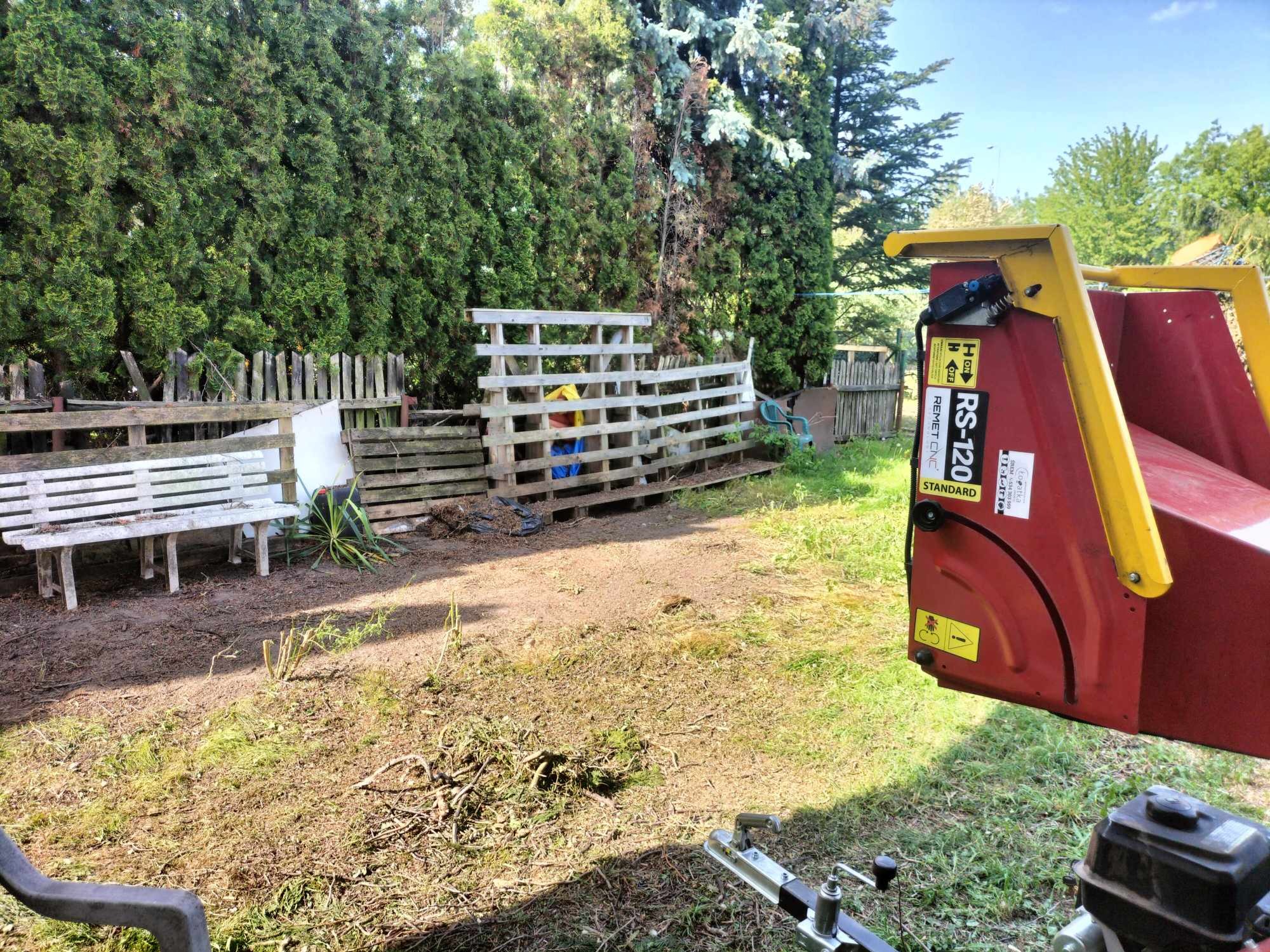 Fragment ogrodu z resztkami po cięciu gałęzi, widoczna maszyna do rozdrabniania gałęzi z żółtymi elementami, w tle drewniany płot i gęste krzewy.