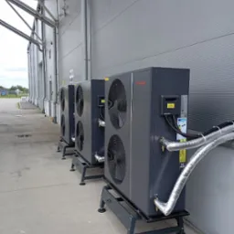 Szereg trzech zainstalowanych urządzeń grzewczych Schellinger na betonowym podłożu, z widocznymi srebrnymi rurami izolacyjnymi i szarą ścianą w tle.