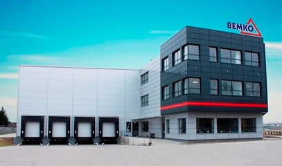 Nowoczesny budynek przemysłowy z logo Bemko pod błękitnym niebem. Szara elewacja, czarne okna i czerwone akcenty. Widoczne rampy załadunkowe.