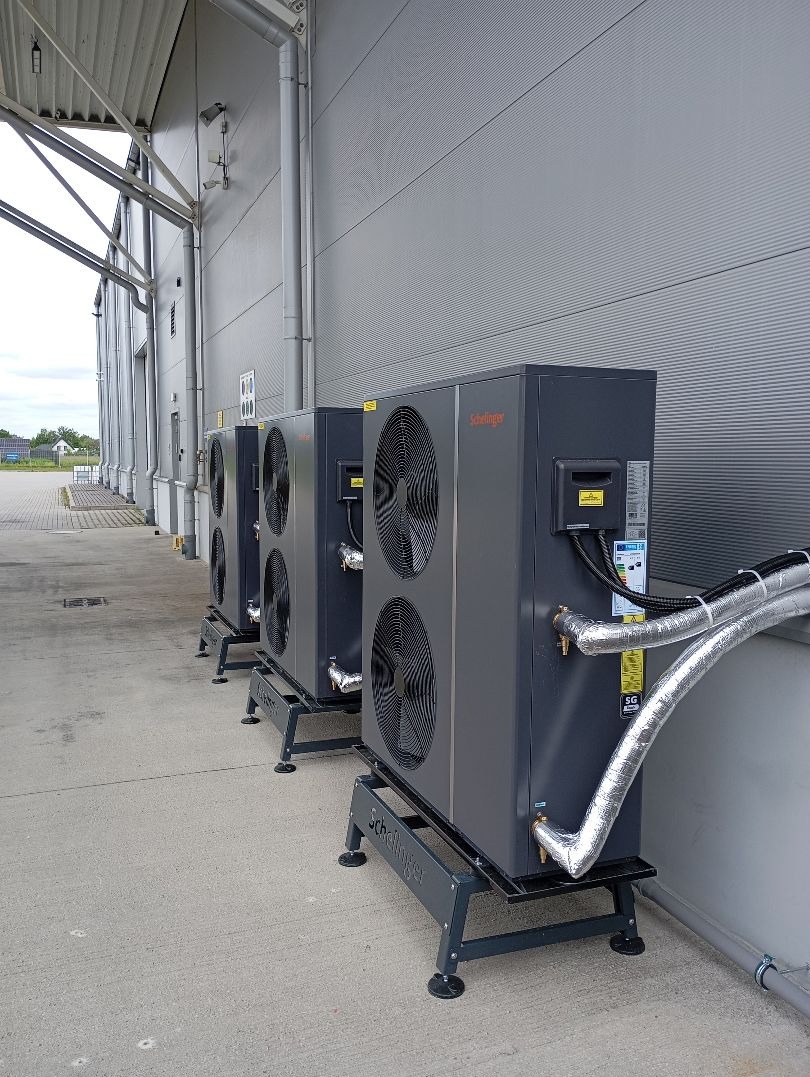 Szereg trzech zainstalowanych urządzeń grzewczych Schellinger na betonowym podłożu, z widocznymi srebrnymi rurami izolacyjnymi i szarą ścianą w tle.