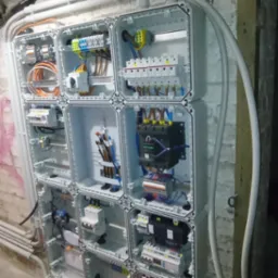 Rozbudowana skrzynka elektryczna z licznymi zabezpieczeniami i przewodami, zamontowana na surowej ścianie z widocznymi rurami instalacyjnymi.