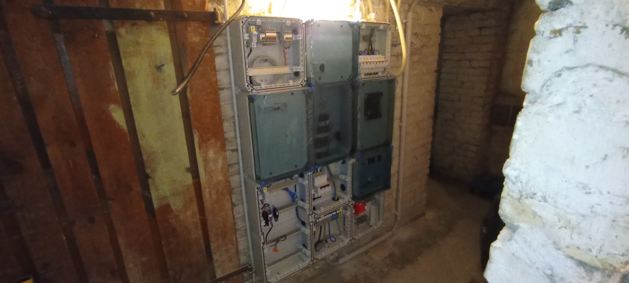 Rozdzielnia elektryczna w piwnicy z drewnianą ścianą i fragmentem muru. Widoczne kable i elementy instalacji. Niektóre skrzynki otwarte.