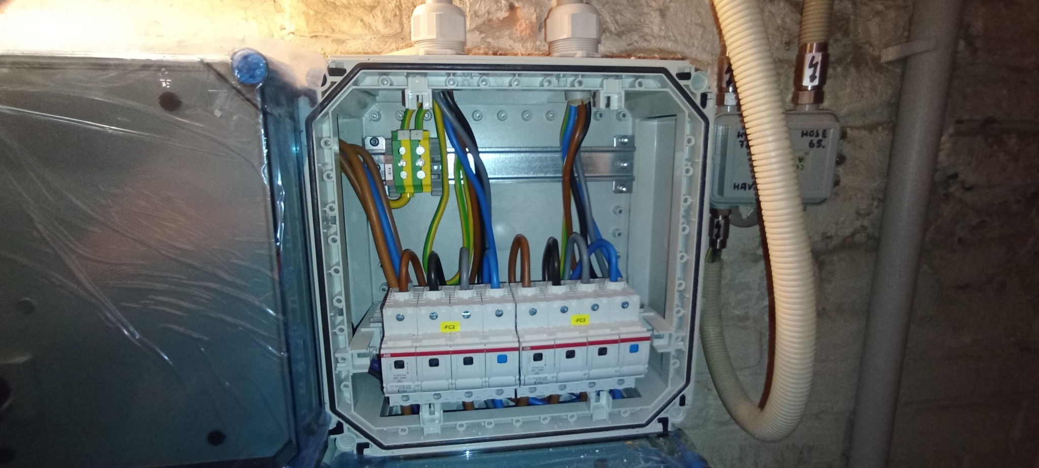 Otwarta skrzynka elektryczna z bezpiecznikami, widoczne przewody w kolorach brązowym, niebieskim, żółto-zielonym. Obok rura karbowana i puszka instalacyjna.