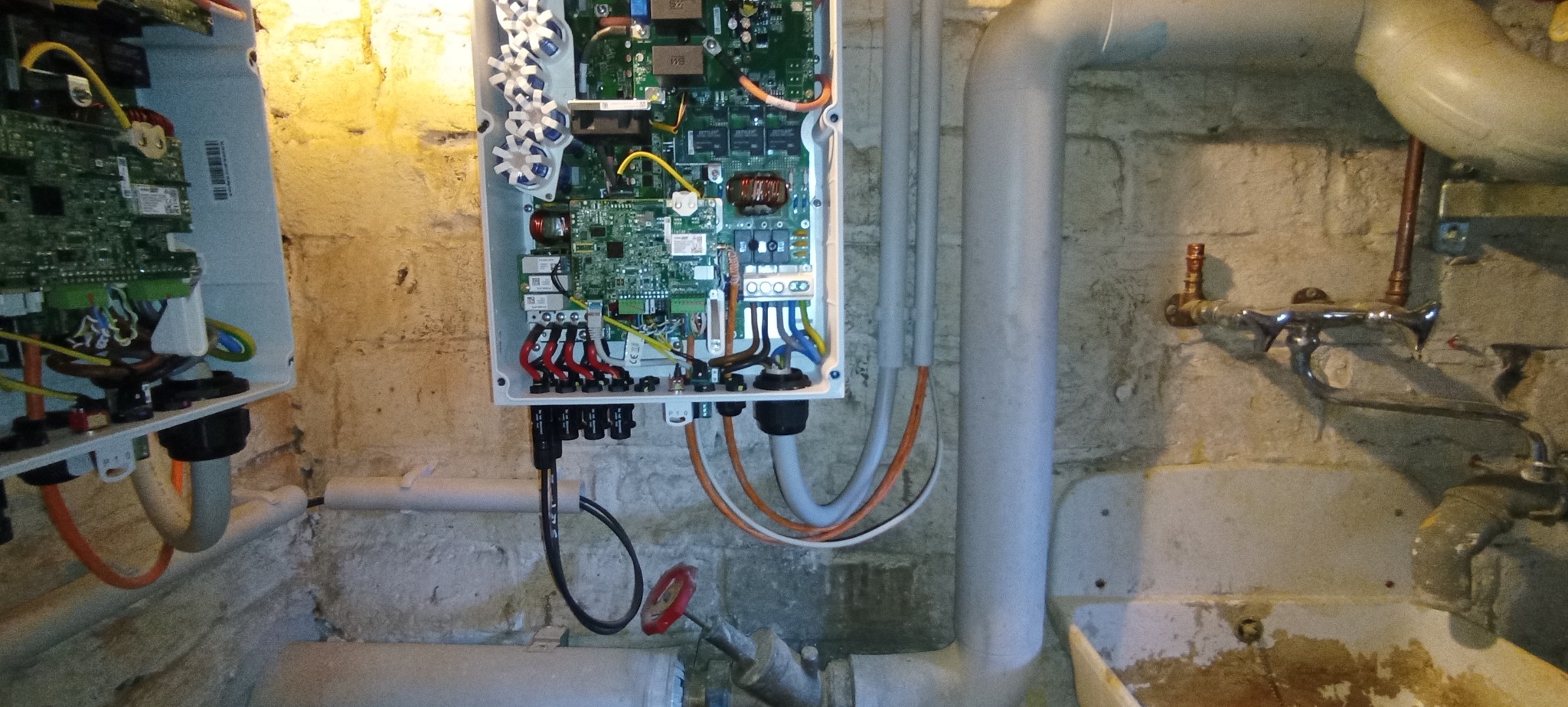 Rozdzielnica elektryczna w piwnicy z widocznymi przewodami i płytkami drukowanymi, na tle ściany z cegły i rur. Widoczne elementy instalacji wodnej.