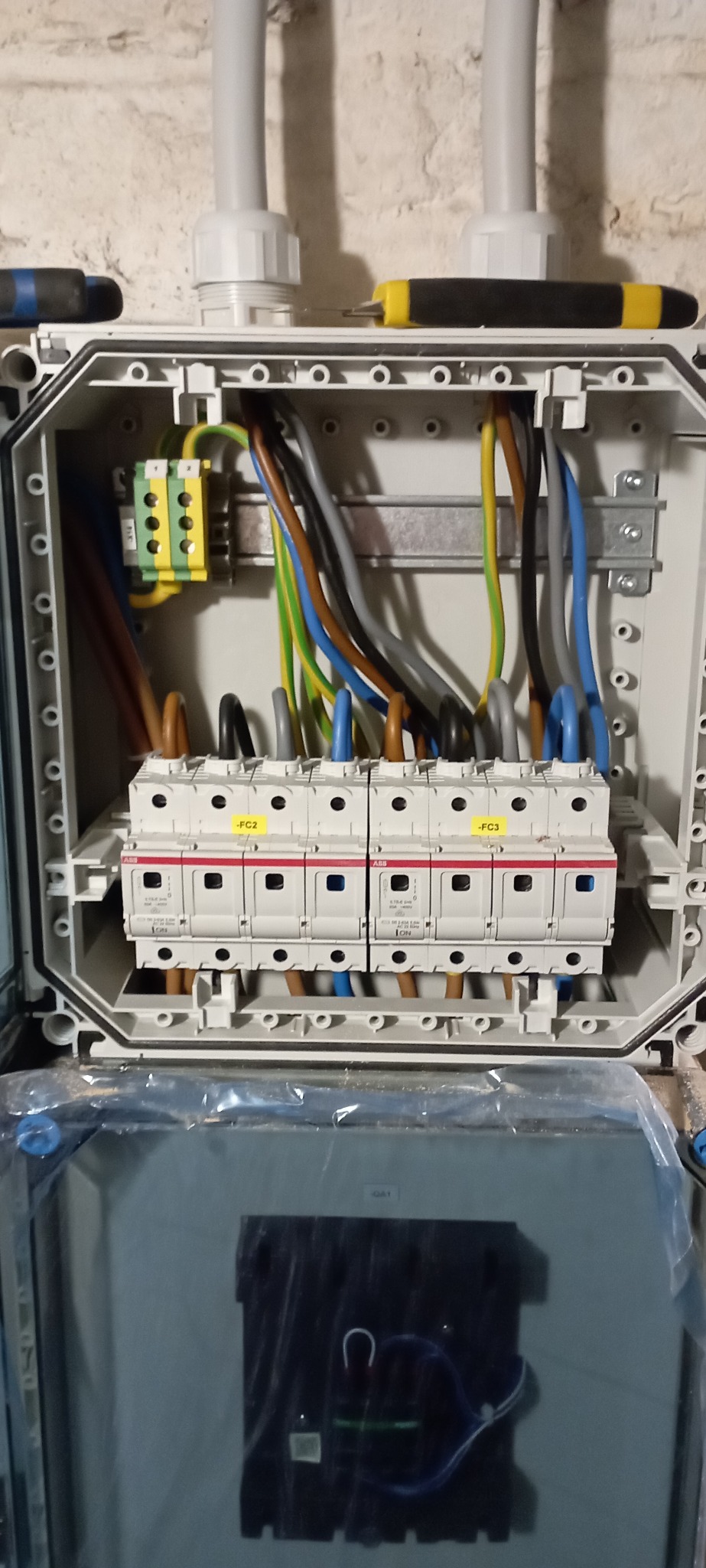 Otwarta skrzynka elektryczna z bezpiecznikami ABB, widoczne kolorowe przewody i listwa zaciskowa. Dwa wkrętaki leżą na obudowie. Biała ściana w tle.