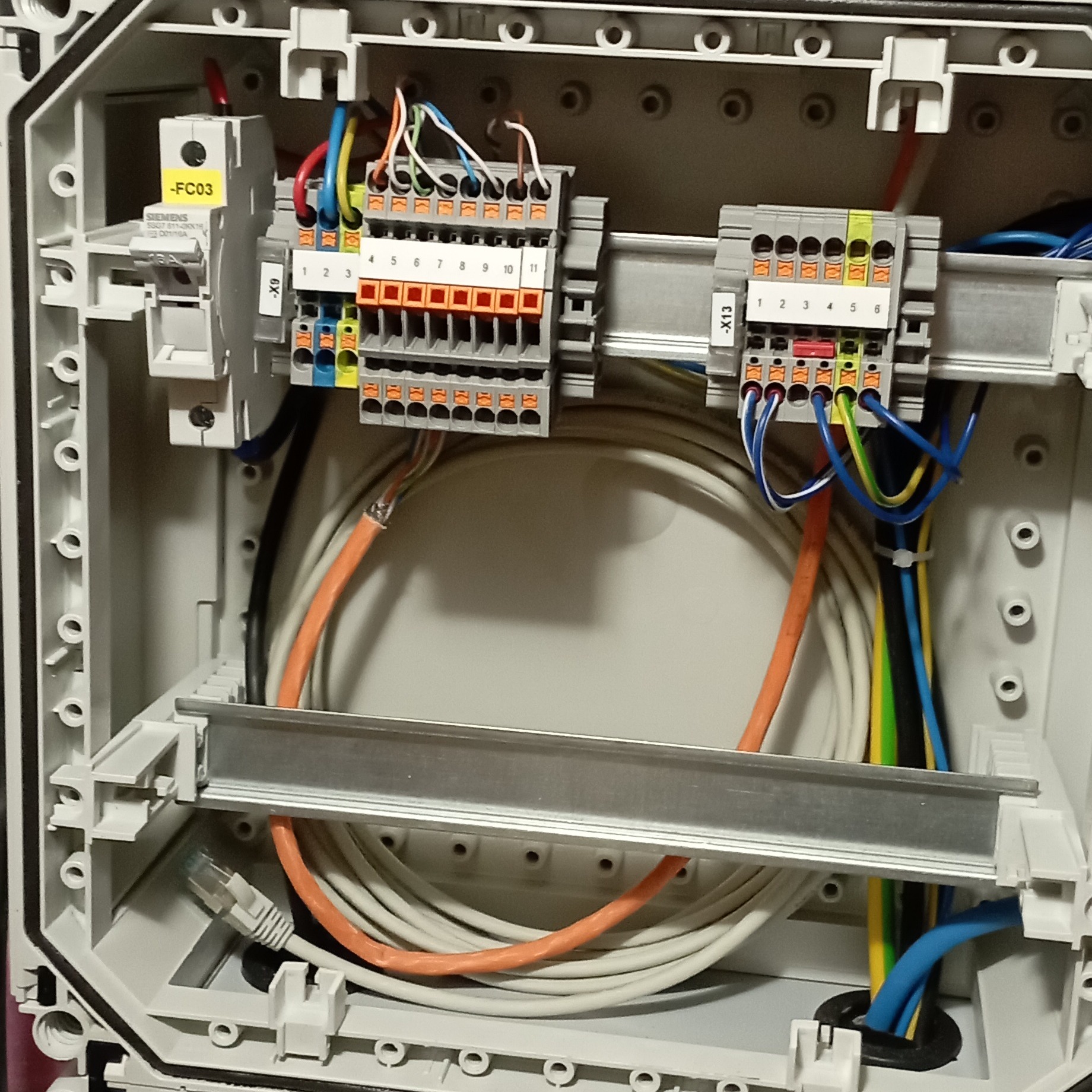 Rozdzielnica elektryczna w obudowie z widocznymi przewodami, złączkami, bezpiecznikiem i kablem Ethernet. Wnętrze skrzynki elektrycznej z okablowaniem i elementami zabezpieczającymi.