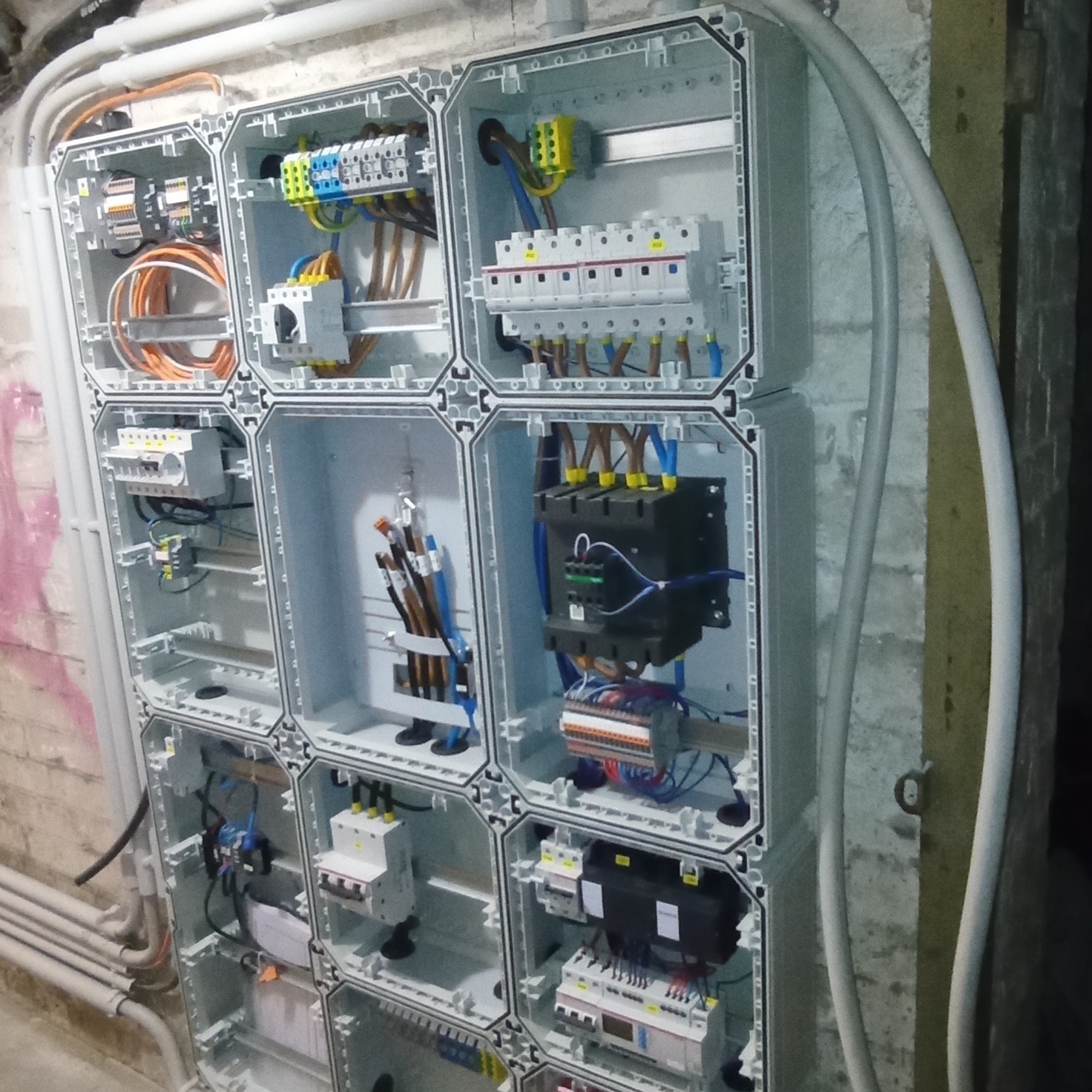 Rozbudowana skrzynka elektryczna z licznymi zabezpieczeniami i przewodami, zamontowana na surowej ścianie z widocznymi rurami instalacyjnymi.