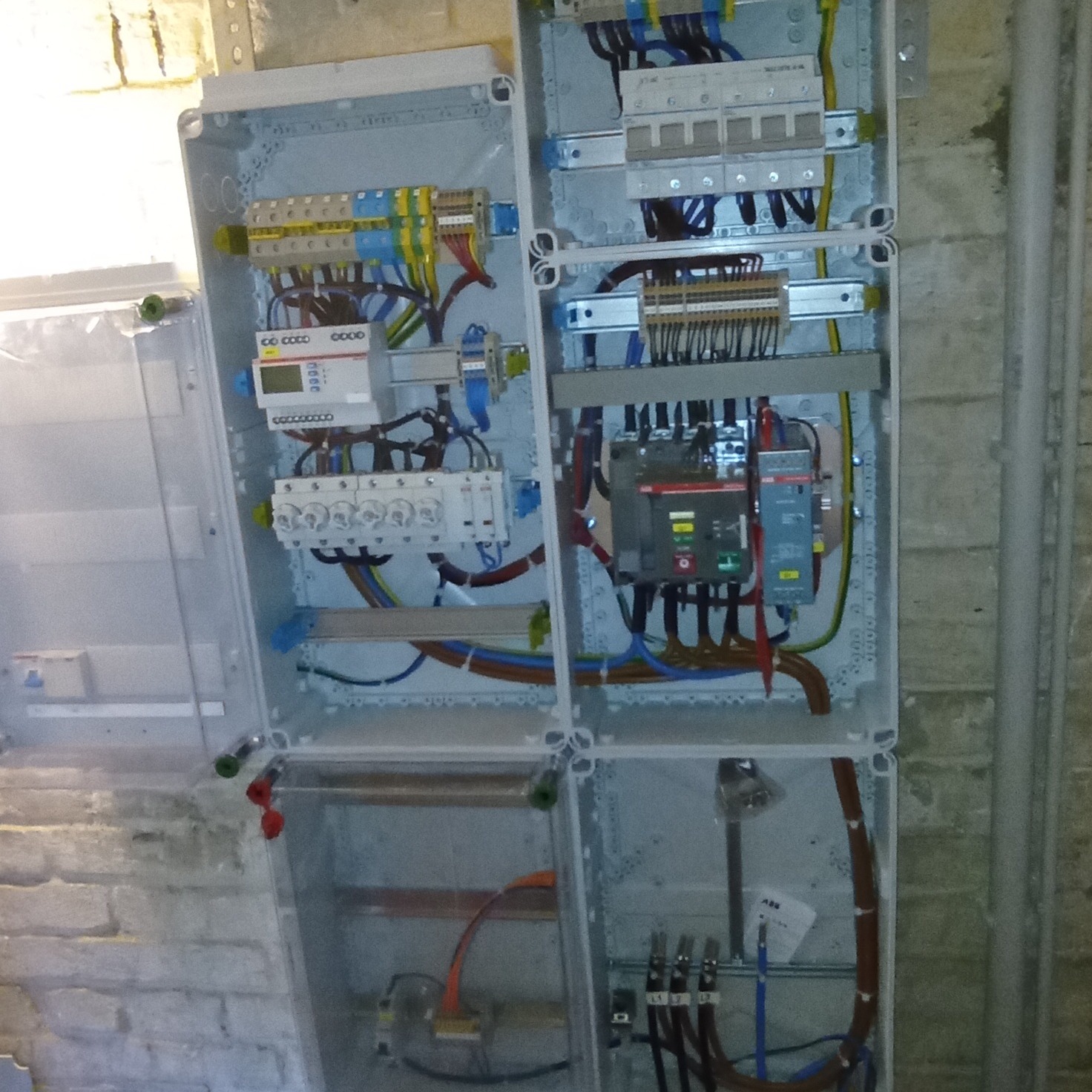 Rozdzielnica elektryczna z otwartą obudową, widoczne zabezpieczenia, przewody i licznik, zamontowana na ścianie z cegły.