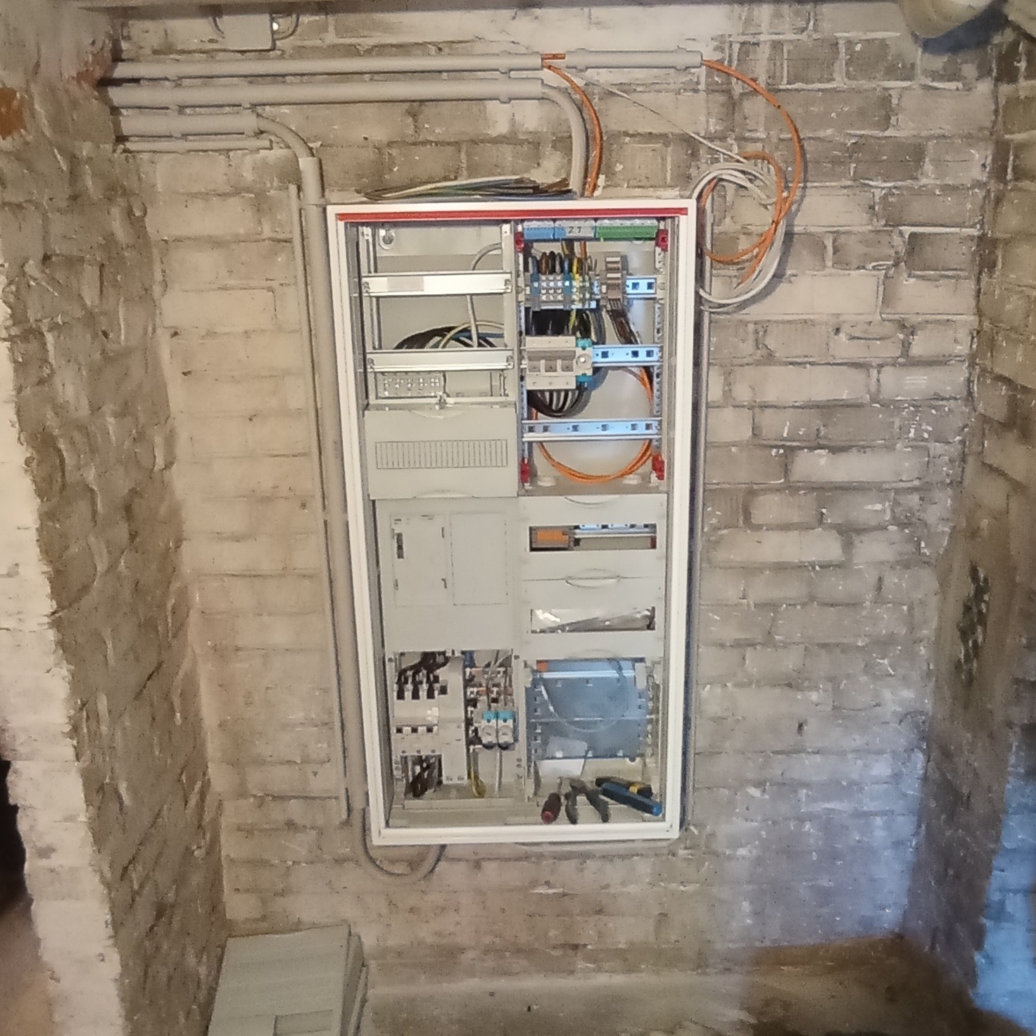 Rozdzielnica elektryczna w białej obudowie na tle surowej ceglanej ściany, widoczne kable i narzędzia. Ujęcie z dołu, instalacja w trakcie montażu.