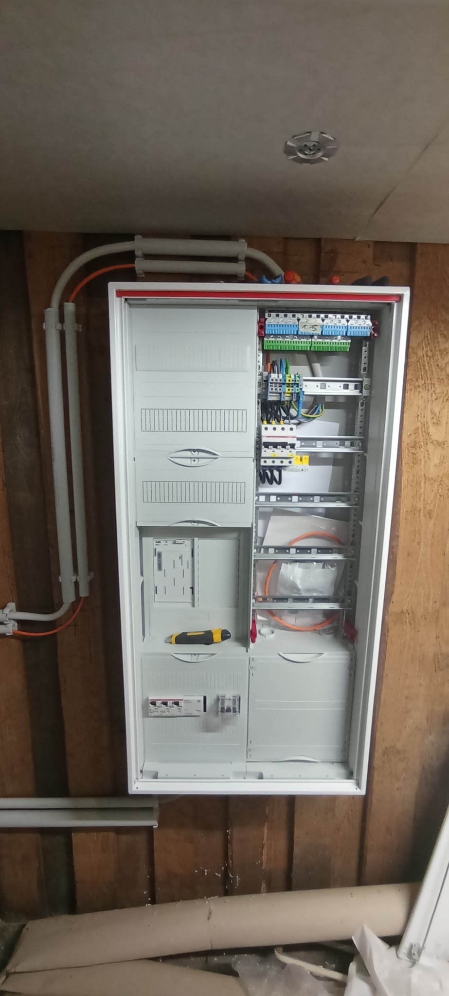 Otwarta, biała skrzynka elektryczna z widocznymi przewodami, bezpiecznikami i narzędziem, zamontowana na drewnianej ścianie. Widoczne rury instalacyjne.