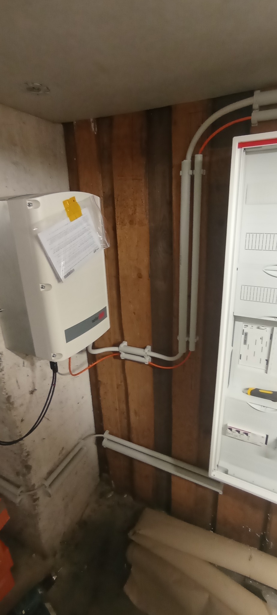 Zamontowana biała skrzynka elektryczna na drewnianej ścianie z widocznymi przewodami i rurkami ochronnymi. Częściowo widoczny panel sterowania.