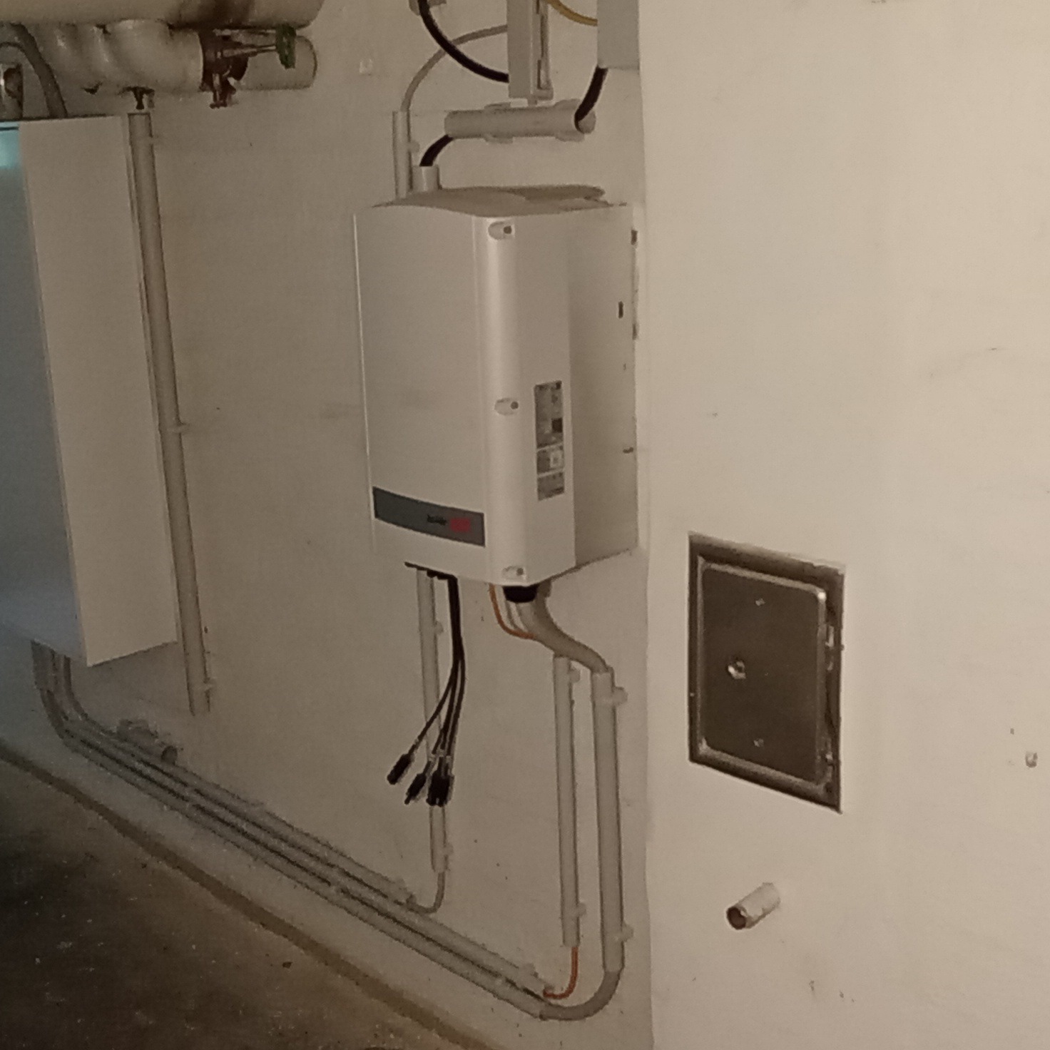 Biały piec elektryczny na ścianie z podłączonymi przewodami. Widoczne rury i skrzynka rewizyjna. Ujęcie z dołu, w piwnicznym pomieszczeniu.