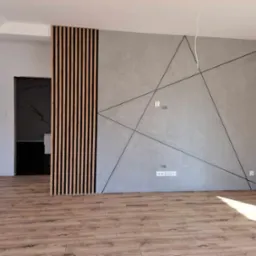 Wykończone wnętrze: szara ściana z geometrycznym wzorem z czarnych linii, drewniane lamele pionowe, podłoga z paneli imitujących drewno, widoczne gniazdka elektryczne.