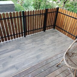WADOM Dominik Walicki - Zadaszony taras z drewnianą balustradą z pionowych desek i czarnymi metalowymi elementami, wyłożony płytkami imitującymi drewno, z wiklinowym fotelem w rogu.