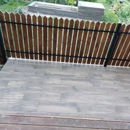 WADOM Dominik Walicki - Nowoczesny taras wyłożony szarymi płytkami imitującymi drewno, z drewnianym ogrodzeniem o pionowych deskach i czarnych metalowych słupkach, widok na zielony ogród z podwyższoną grządką.