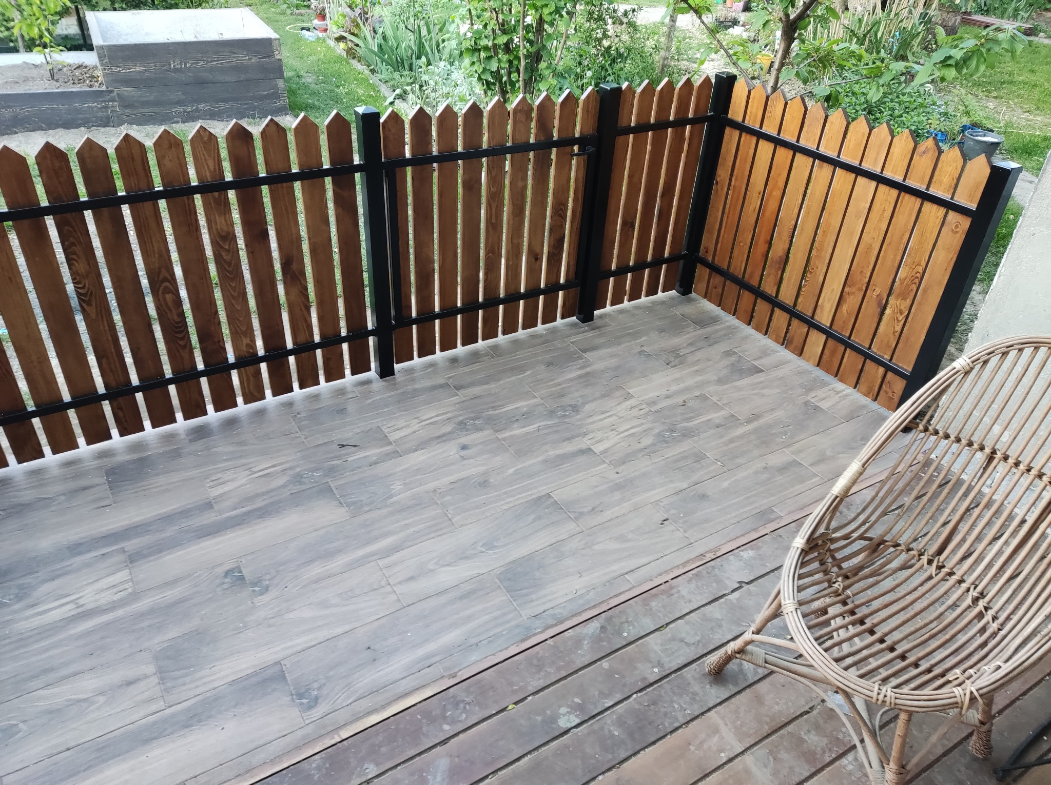 Zadaszony taras z drewnianą balustradą z pionowych desek i czarnymi metalowymi elementami, wyłożony płytkami imitującymi drewno, z wiklinowym fotelem w rogu.