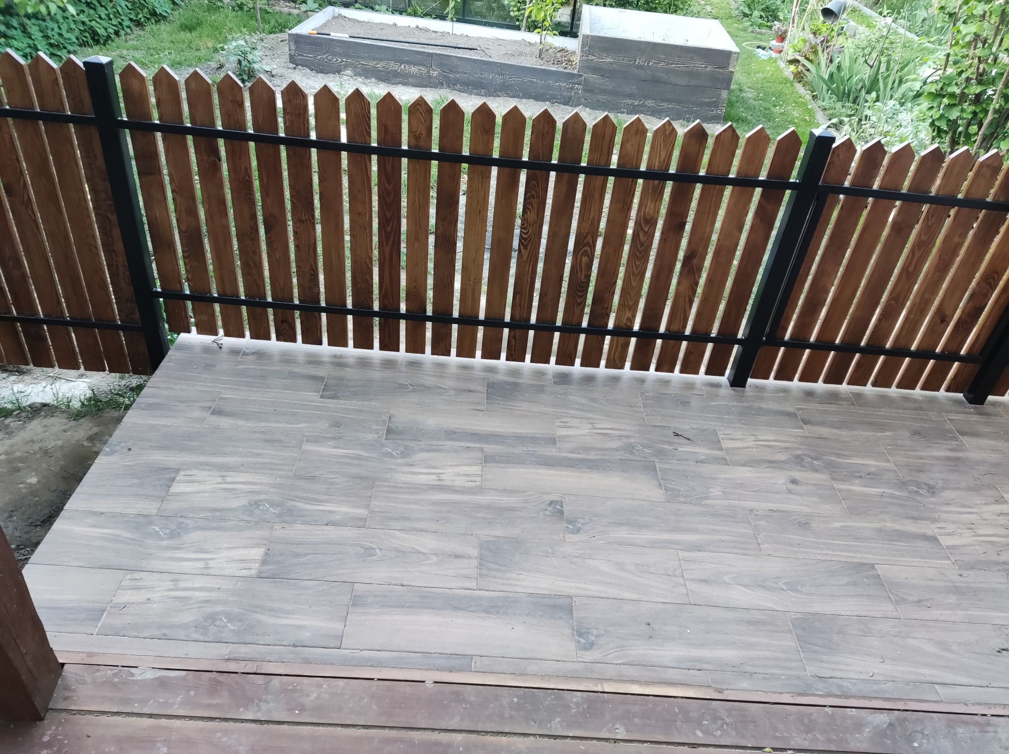 Nowoczesny taras wyłożony szarymi płytkami imitującymi drewno, z drewnianym ogrodzeniem o pionowych deskach i czarnych metalowych słupkach, widok na zielony ogród z podwyższoną grządką.