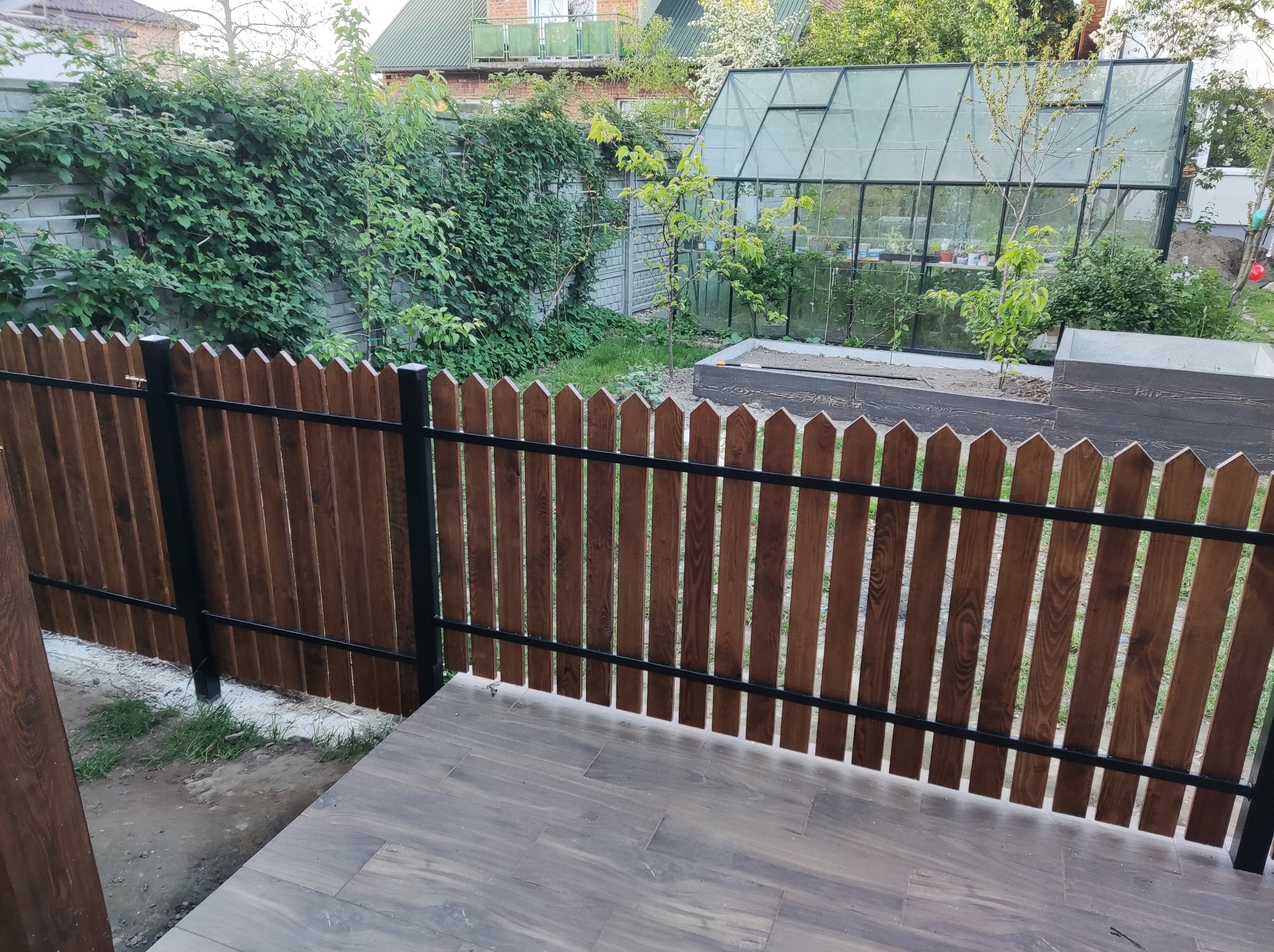 Nowoczesne ogrodzenie drewniane z czarnymi metalowymi słupkami, oddzielające taras z płytek imitujących drewno od zielonego ogrodu z szklarnią w tle.