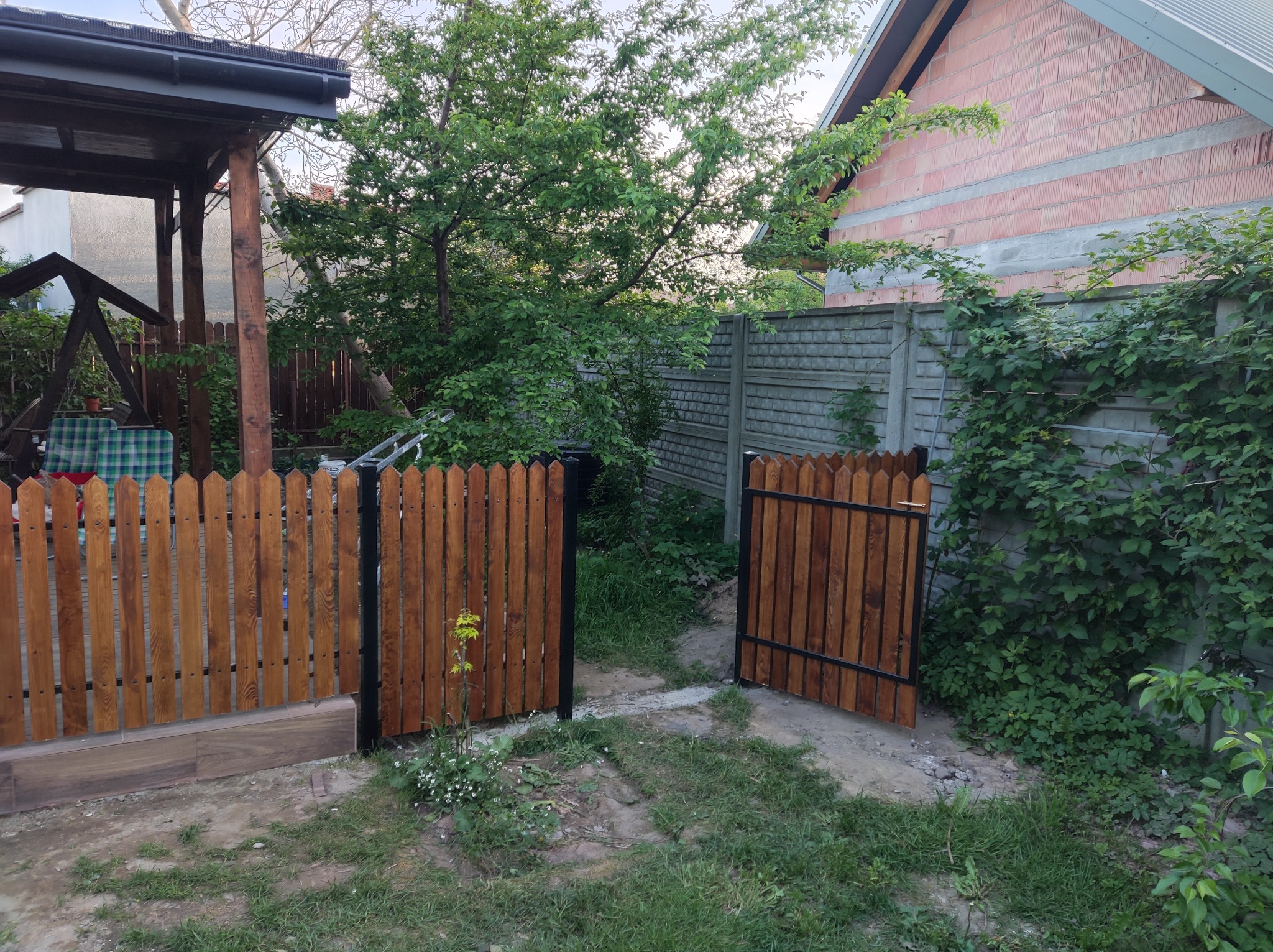 Nowo zainstalowane drewniane ogrodzenie z furtką, pomalowane na brązowo, z czarnymi metalowymi ramami, oddzielające ogród od posesji z betonowym ogrodzeniem i budynkiem w trakcie budowy.
