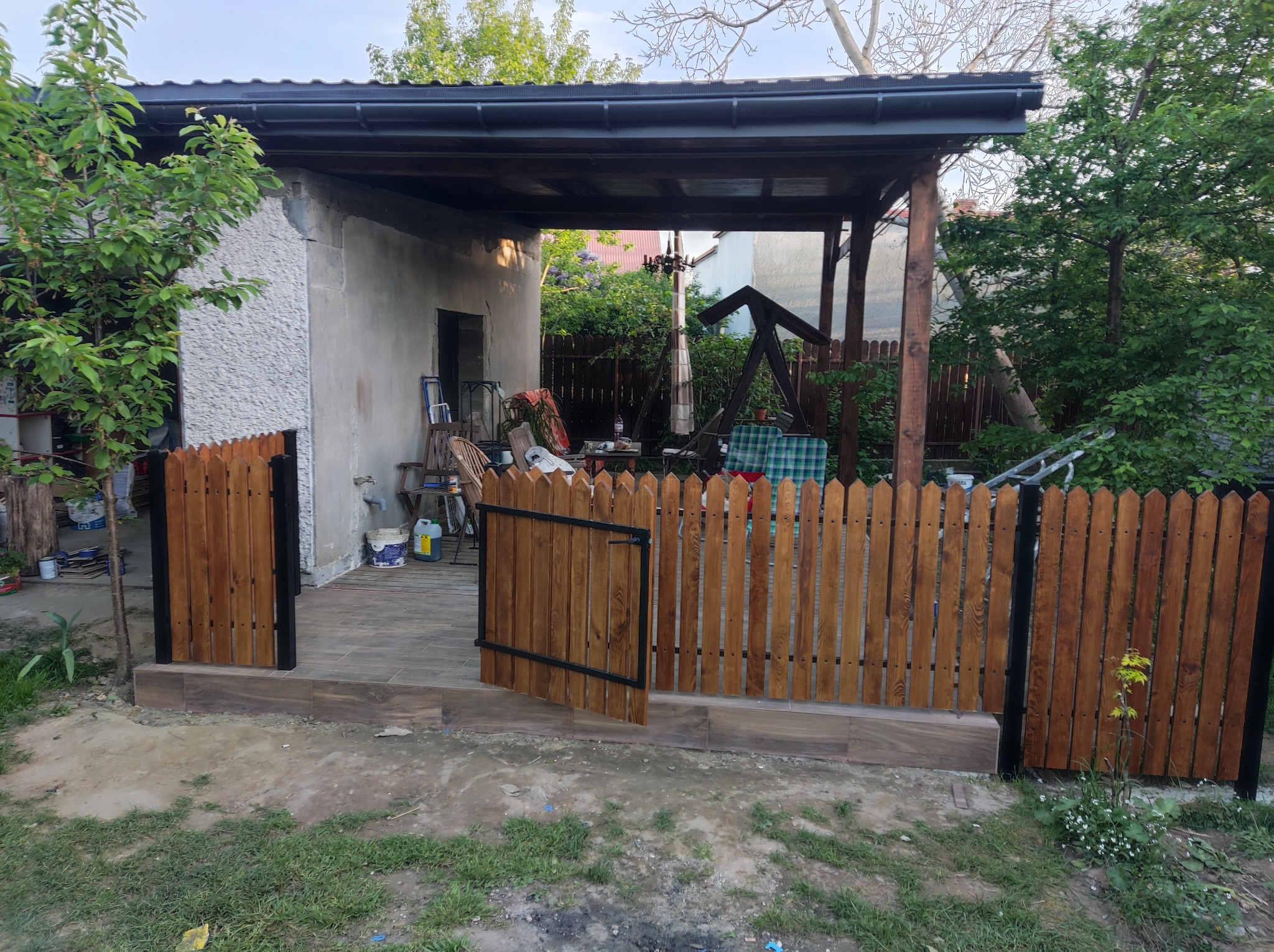 Nowo postawione drewniane ogrodzenie z furtką, pomalowane na brązowo z czarnymi elementami, oddzielające taras z płytkami od trawnika.
