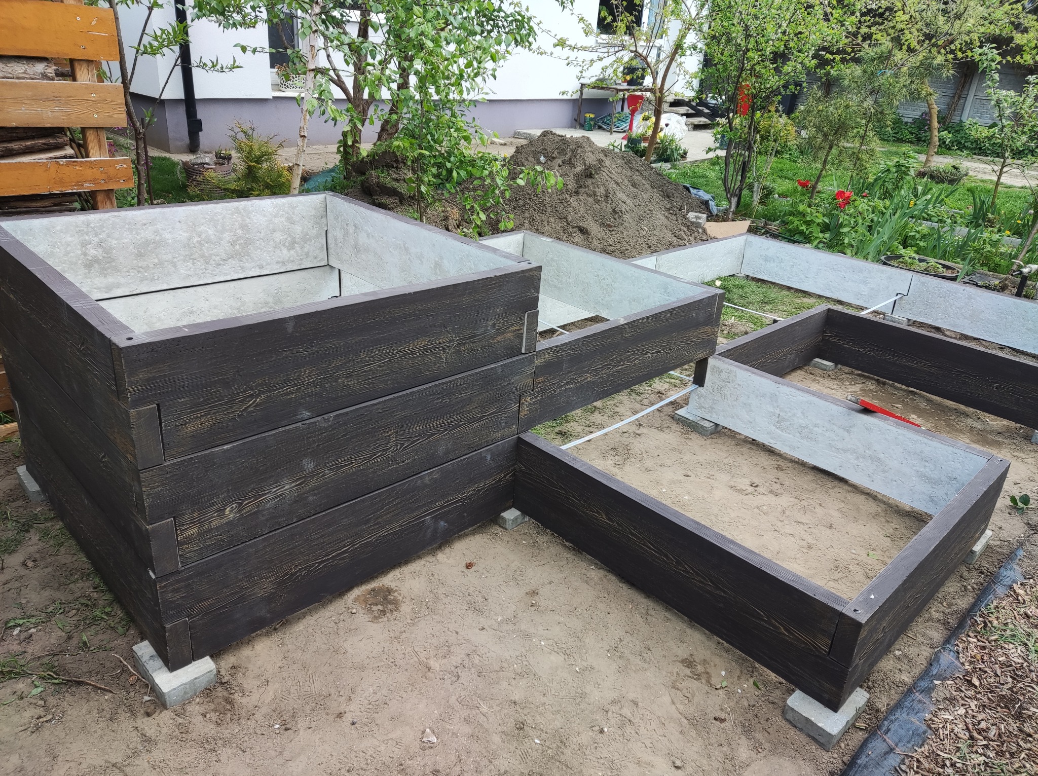Wielopoziomowa konstrukcja z ciemnego drewna imitującego deski, tworząca podwyższone rabaty ogrodowe, ustawiona na betonowych bloczkach w ogrodzie.