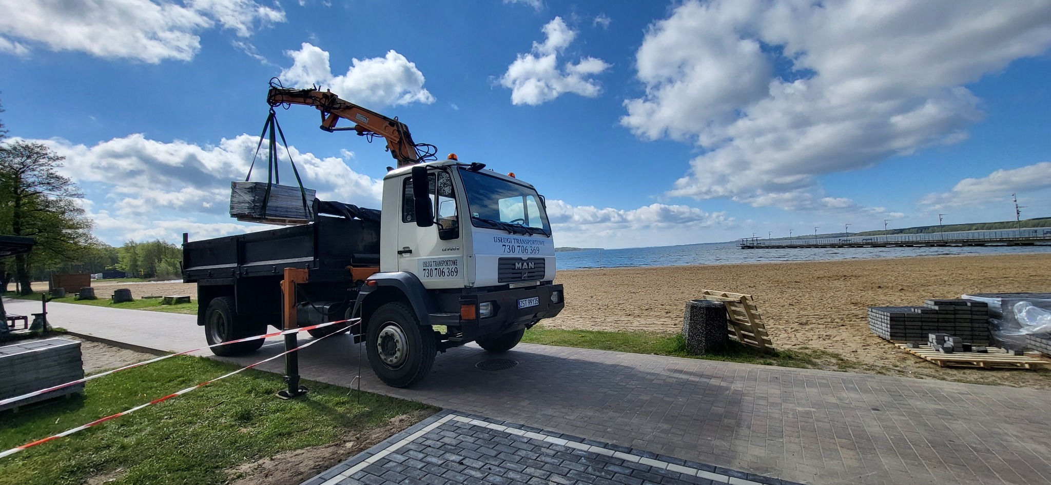 Ciężarówka MAN z dźwigiem, załadowana kostką brukową, na tle plaży i jeziora pod błękitnym niebem z chmurami.