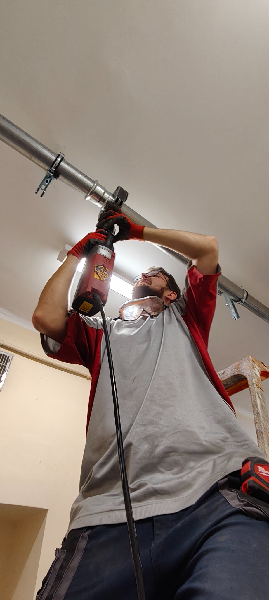 Pracownik w okularach ochronnych, w trakcie montażu rury instalacyjnej pod sufitem, używając specjalistycznego narzędzia. Widoczna drabina i elementy instalacji.
