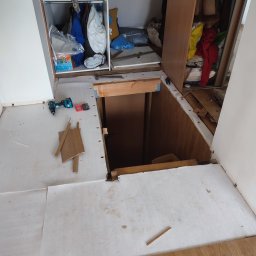 REMONTPRO USŁUGI REMONTOWO - BUDOWLANE SZYMON BRZĘCZKOWSKI - Fragment podłogi w trakcie demontażu, widoczny otwór w podłodze, wiertarka i drobne elementy drewniane na białej powierzchni, otwarte szafy w tle.