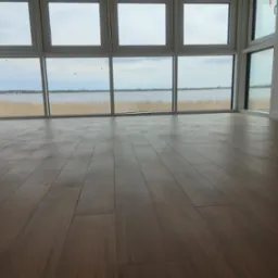 Nowo ułożone, ciemnobrązowe panele podłogowe w jasnym pokoju z dużymi oknami z widokiem na jezioro i trzciny; widoczne detale łączeń paneli.