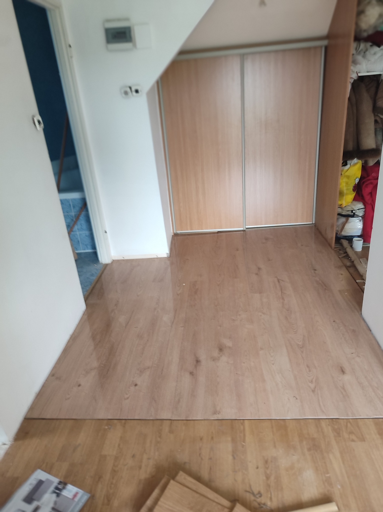 Fragment korytarza z nowo ułożonymi panelami podłogowymi w odcieniach jasnego drewna, widoczne przejście do łazienki z niebieskimi płytkami oraz fragment szafy przesuwnej z jasnego drewna.