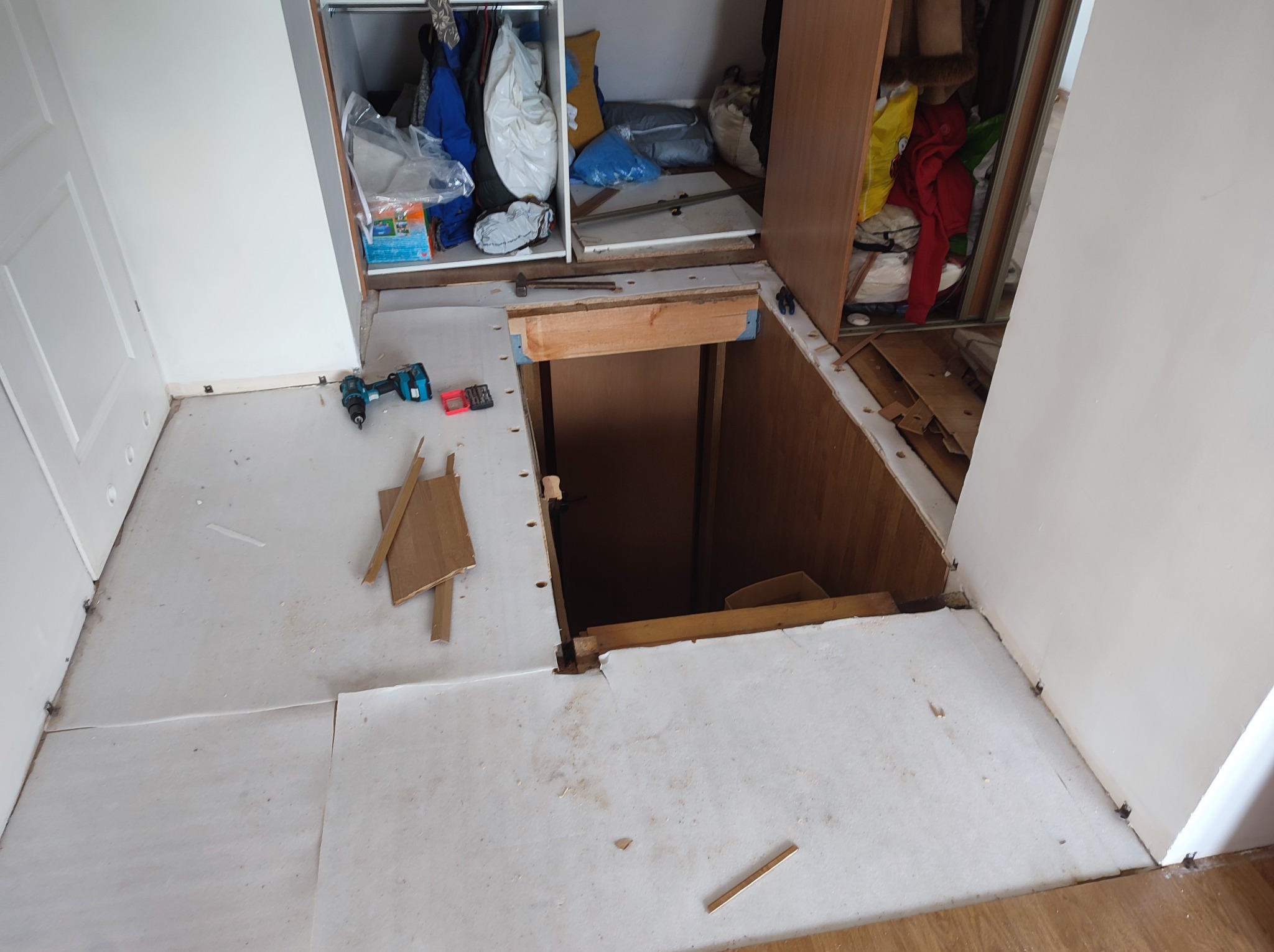 Fragment podłogi w trakcie demontażu, widoczny otwór w podłodze, wiertarka i drobne elementy drewniane na białej powierzchni, otwarte szafy w tle.