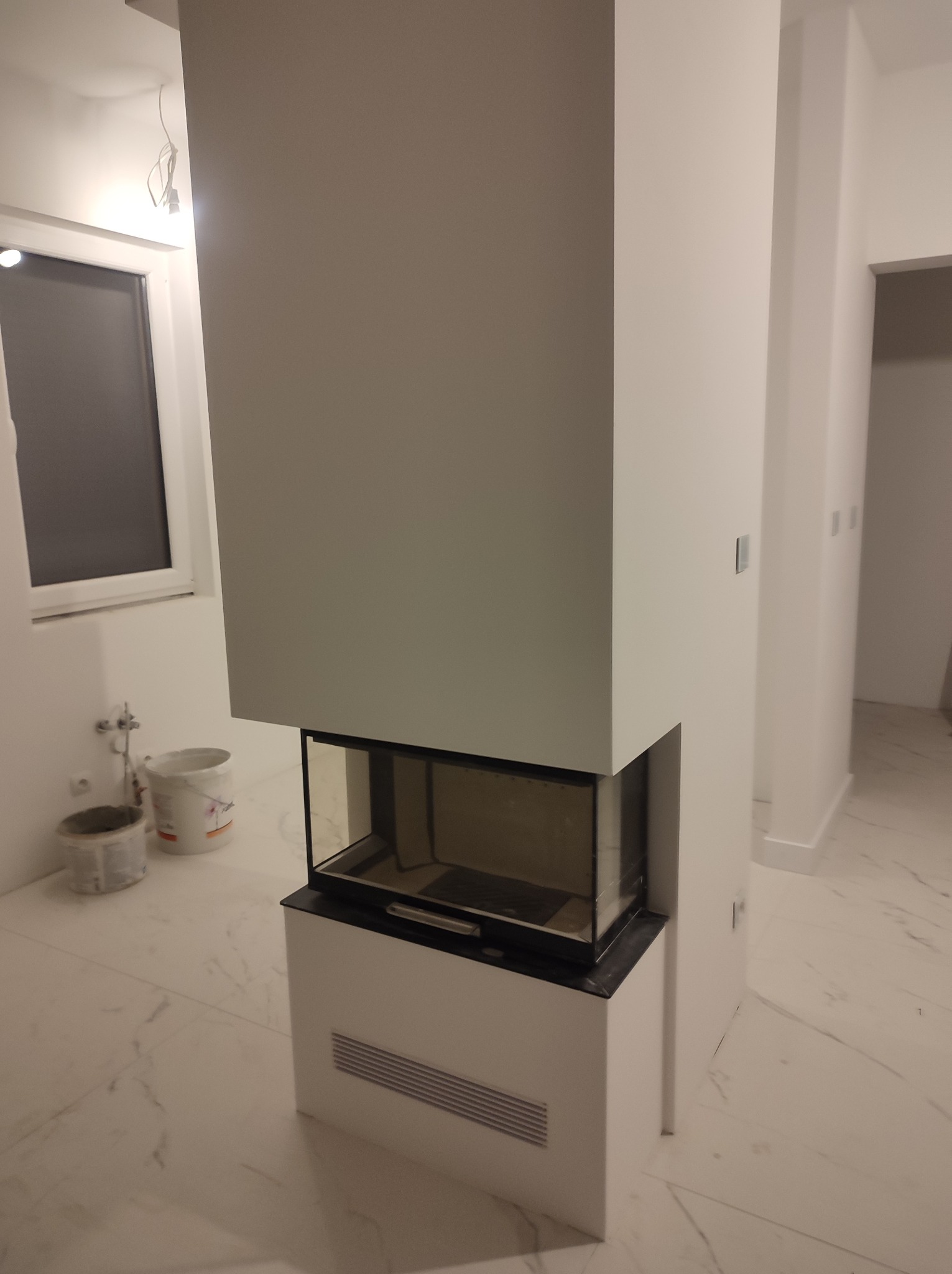 Nowoczesny, biały kominek z szybą w trakcie montażu w Kamieniu Pomorskim, widoczne wiaderka z farbą i marmurowe płytki podłogowe.