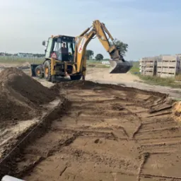 Żółta koparko-ładowarka CAT podczas prac ziemnych na placu budowy, widoczne wytyczone obrysy fundamentów i rolka geowłókniny, w tle ułożone palety z kostką brukową.
