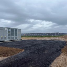 EKOPLANET Sp. z o.o. - Szary kontener z instalacją elektryczną na terenie farmy fotowoltaicznej, z wysypanym świeżym, czarnym kruszywem, panele słoneczne w tle, pochmurne niebo.