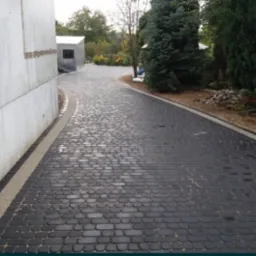 Czarna kostka brukowa ułożona na podjeździe w kształcie łuku, z jasnoszarą obwódką i budynkiem po lewej stronie oraz zielenią po prawej.