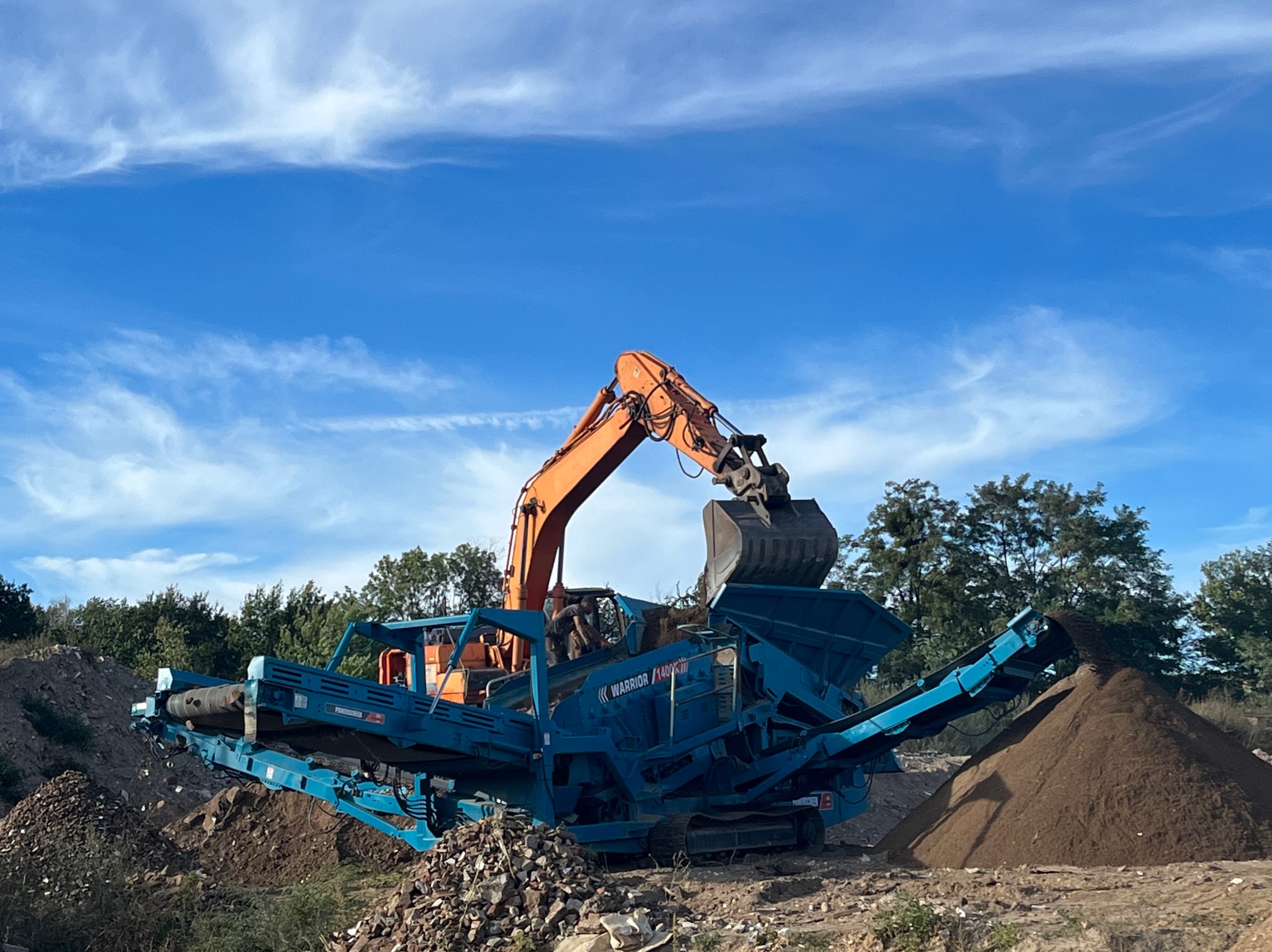 Pomarańczowa koparka ładuje ziemię do niebieskiej przesiewarki Warrior 1400X, formując pryzmę ziemi na tle błękitnego nieba i zieleni.