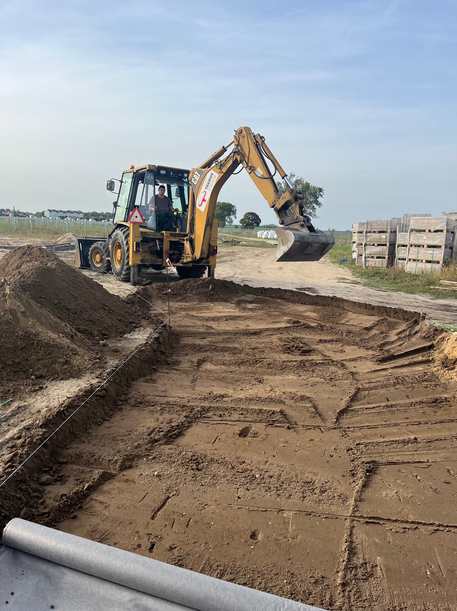 Żółta koparko-ładowarka CAT podczas prac ziemnych na placu budowy, widoczne wytyczone obrysy fundamentów i rolka geowłókniny, w tle ułożone palety z kostką brukową.