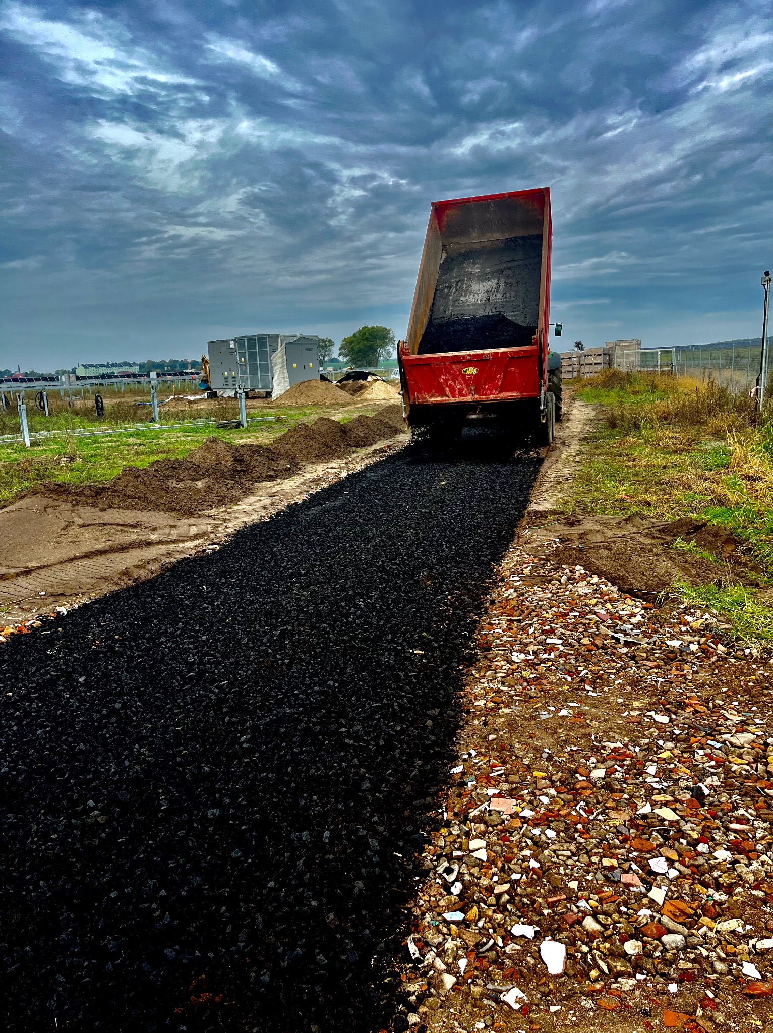 Czerwona wywrotka wysypuje czarny gruz na przygotowany teren pod budowę drogi, widoczne fragmenty starej nawierzchni i odległe budynki oraz instalacje.