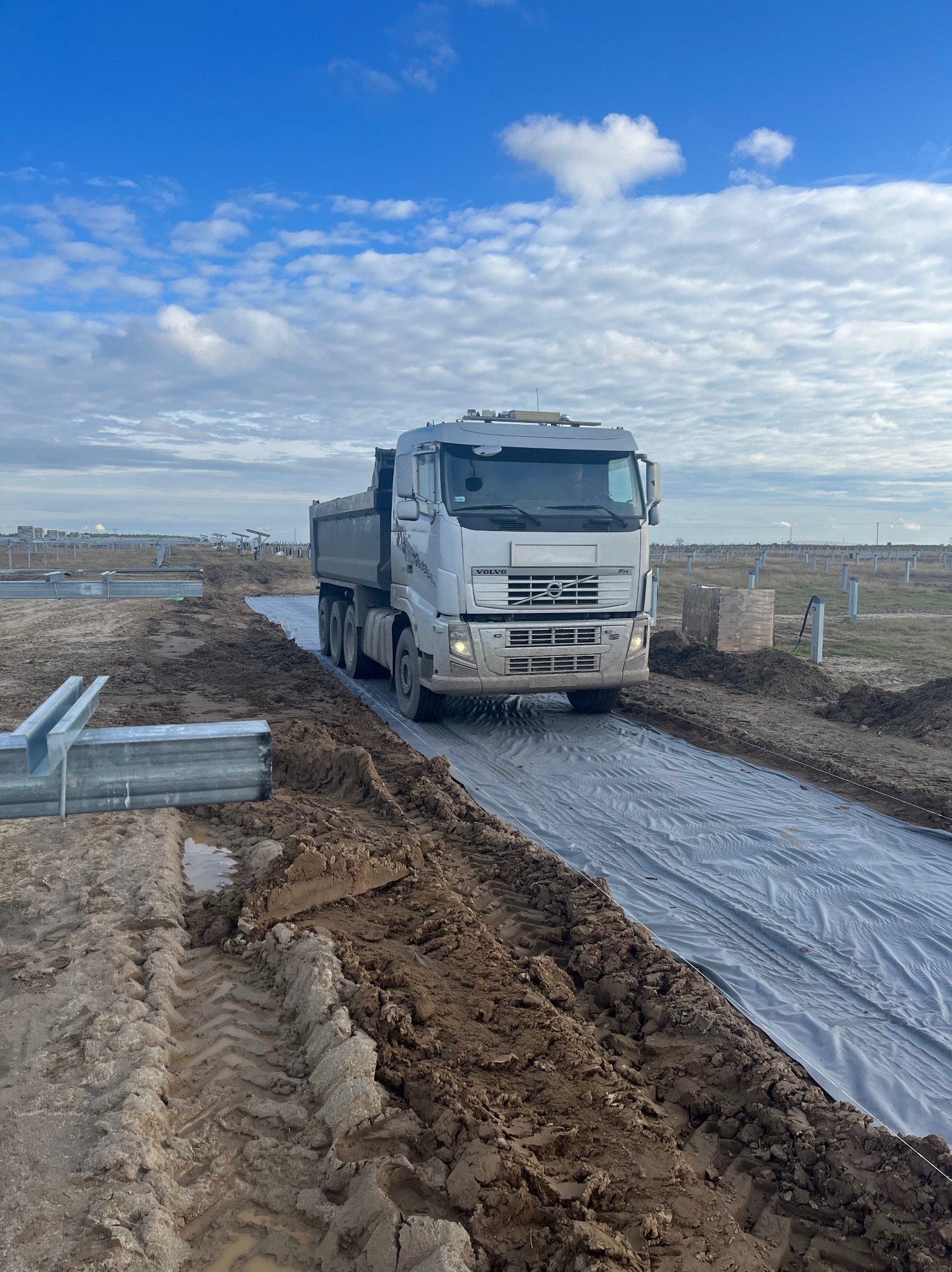 Ciężarówka Volvo FH na placu budowy, jadąca po szarej folii, w tle widoczne metalowe konstrukcje i słupy, błękitne niebo z chmurami.